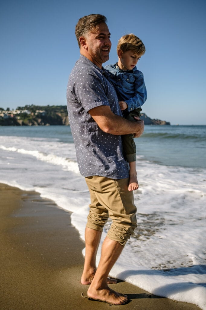 Dad holds son on beach while water washes over his bare feet