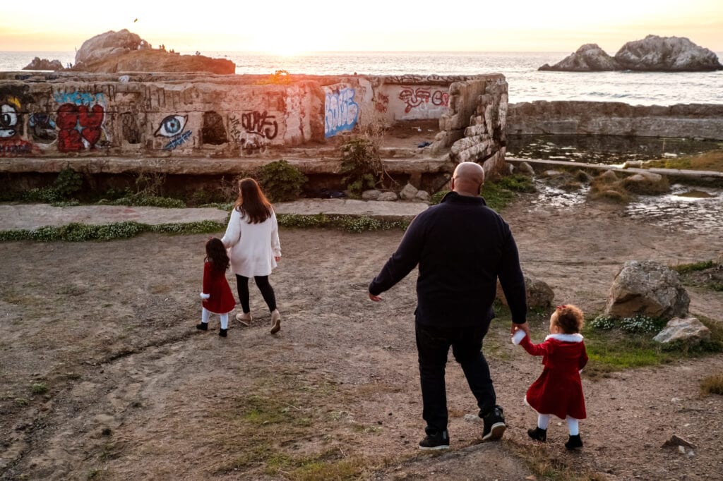 Mom and dad each hold one kids hand as they walk towards the Sutro Baths ruins at sunset.