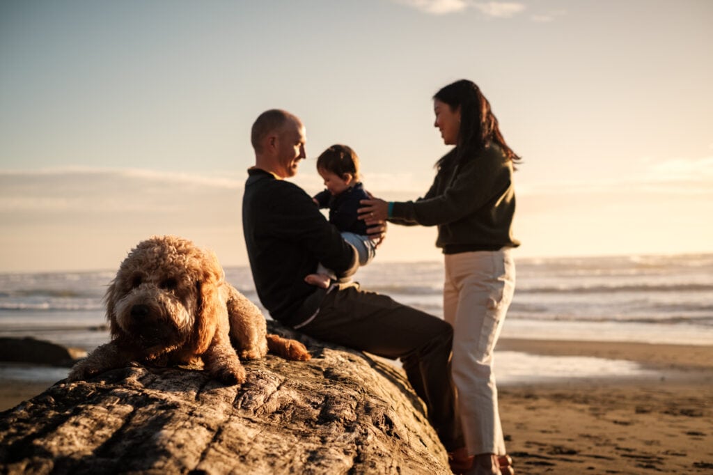 Family dog sits on a log in the foreground of a young couple and their baby.