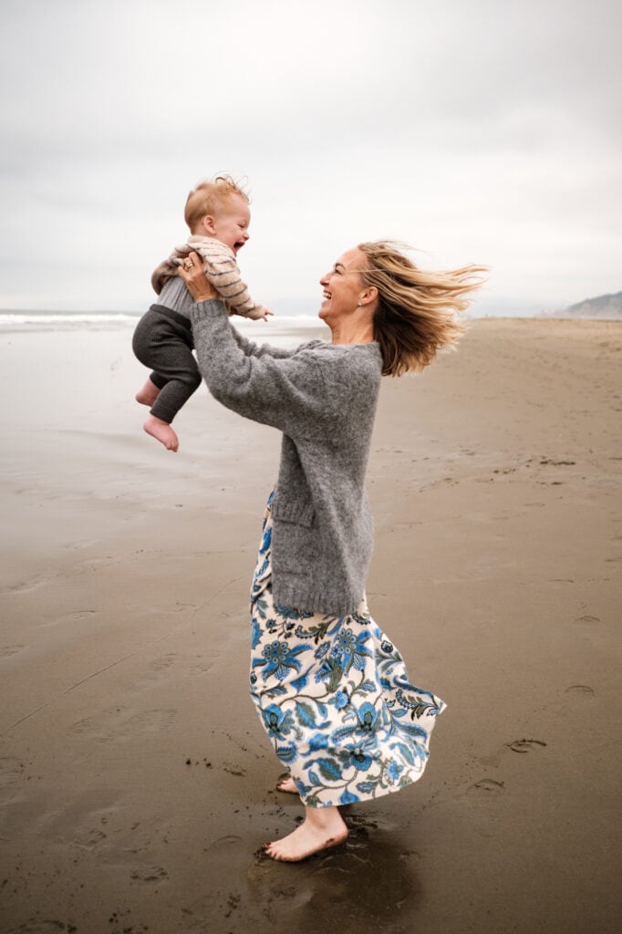 Woman in flowy dress holds her toddler above her on the beach.