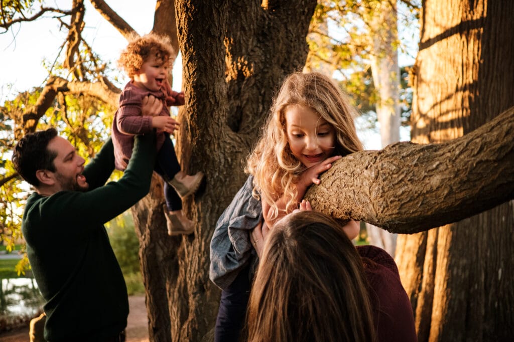 Daughters smile and laugh as parents help them climb a tree.