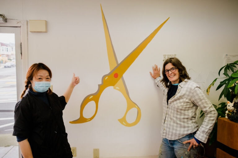 Artist and small business owner stand in front of scissors mural