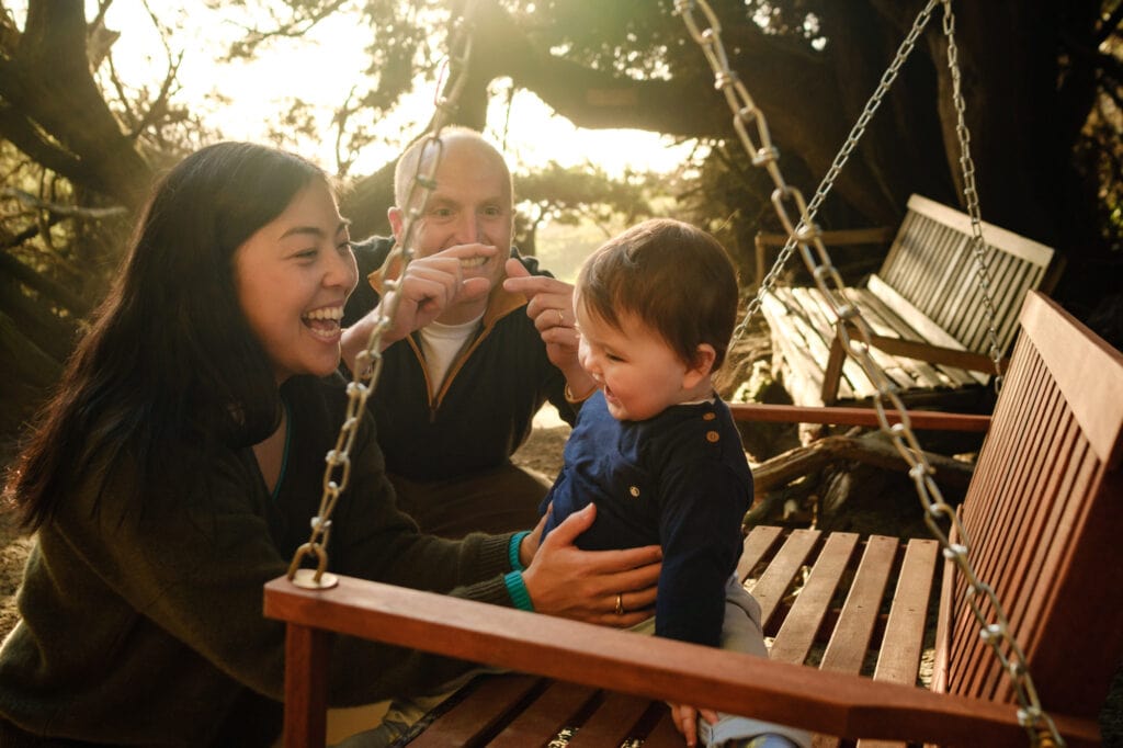 Infant laughs with parents while sitting on a bench in a dense tree nook.