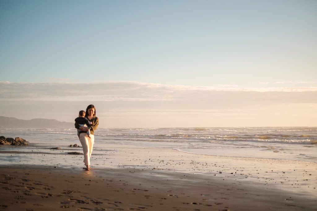 Woman with infant walks down empty beach in sunset light.