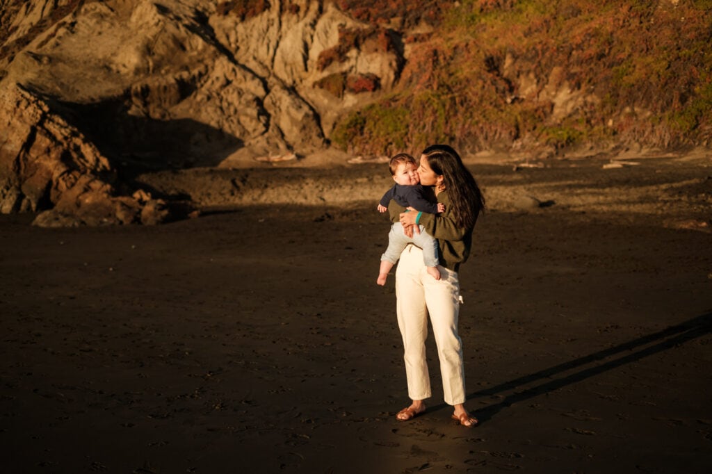 Woman kisses infant while standing on beach in front of dark sandstone cliffs.