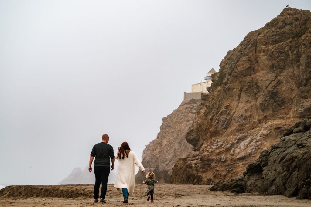 Mom dad and daughter walk on foggy beach below San Francisco Cliff House