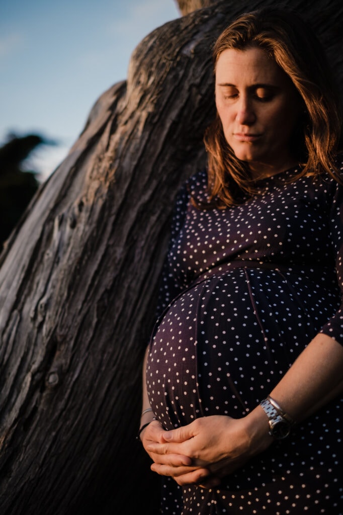 Pregnant woman leans against cypress tree.