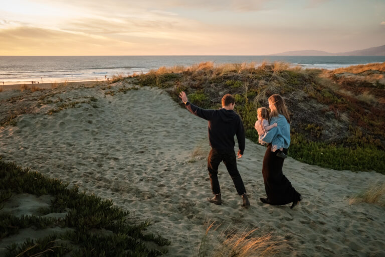 Dad points towards ocean as mom holds daughter and walks up sand dune towards sunset.