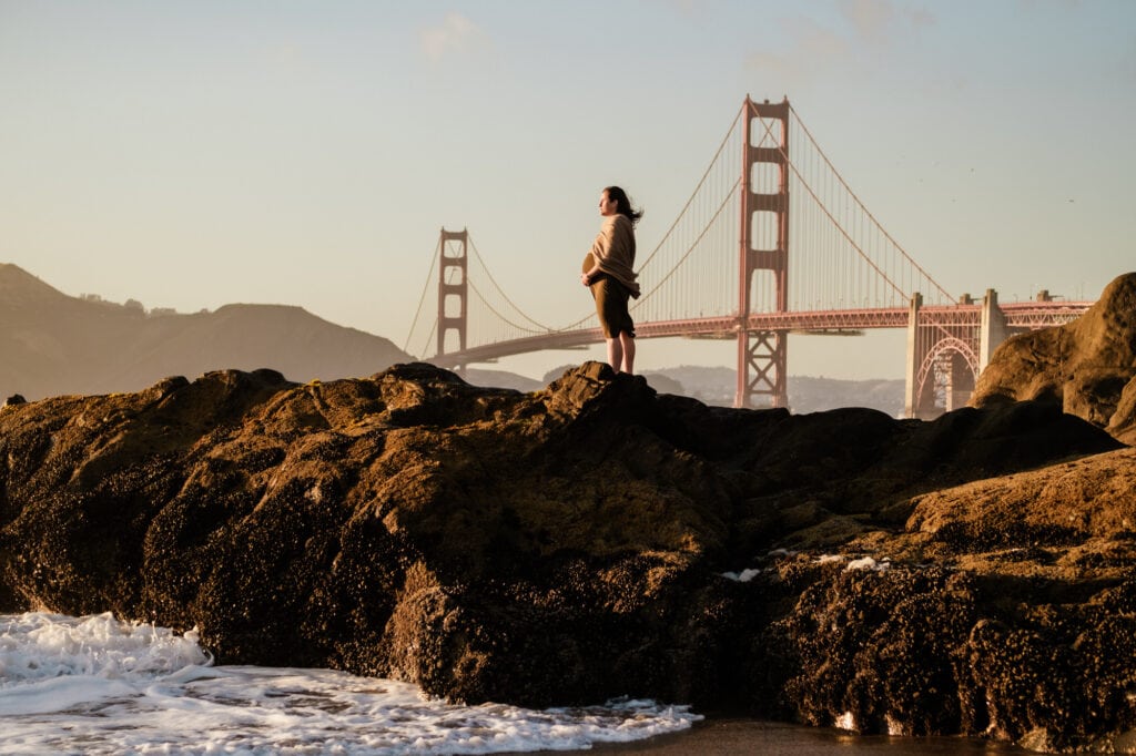 Pregnant woman stands on rocks and looks into sunset in front of the Golden Gate Bridge.