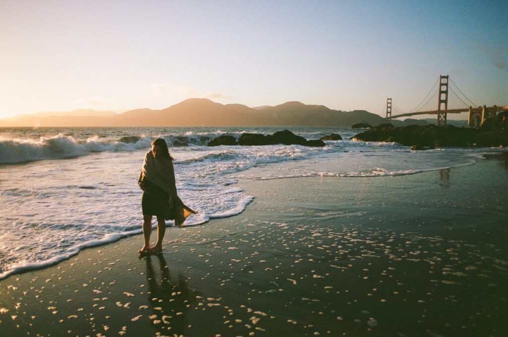 Pregnant woman walks barefoot at the water's edge in sunset light with the Golden Gate Bridge behind