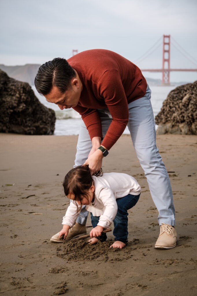 Dad holds daughter's hair as she plays in the sand. Golden Gate Bridge behind.