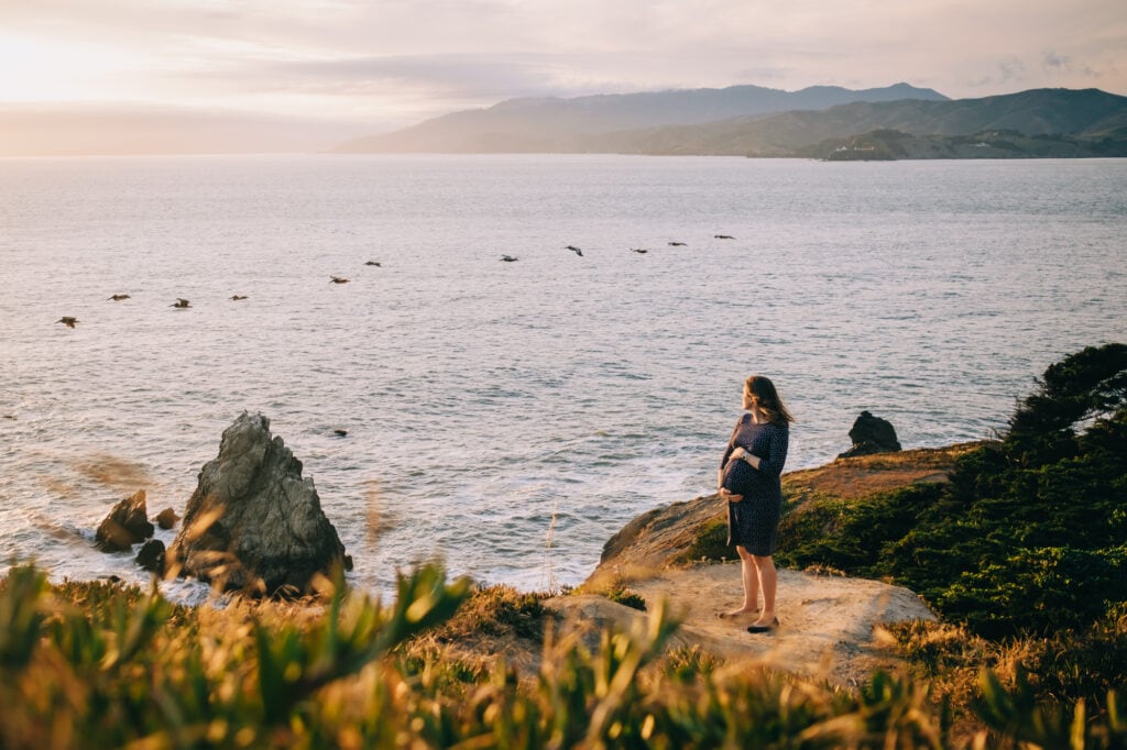 Pregnant woman stands atop sea cliffs and watches birds fly above the ocean.