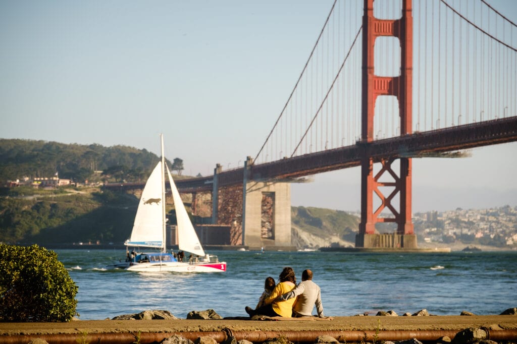 Family of three sit and watch a boat sail under the Golden Gate Bridge.
