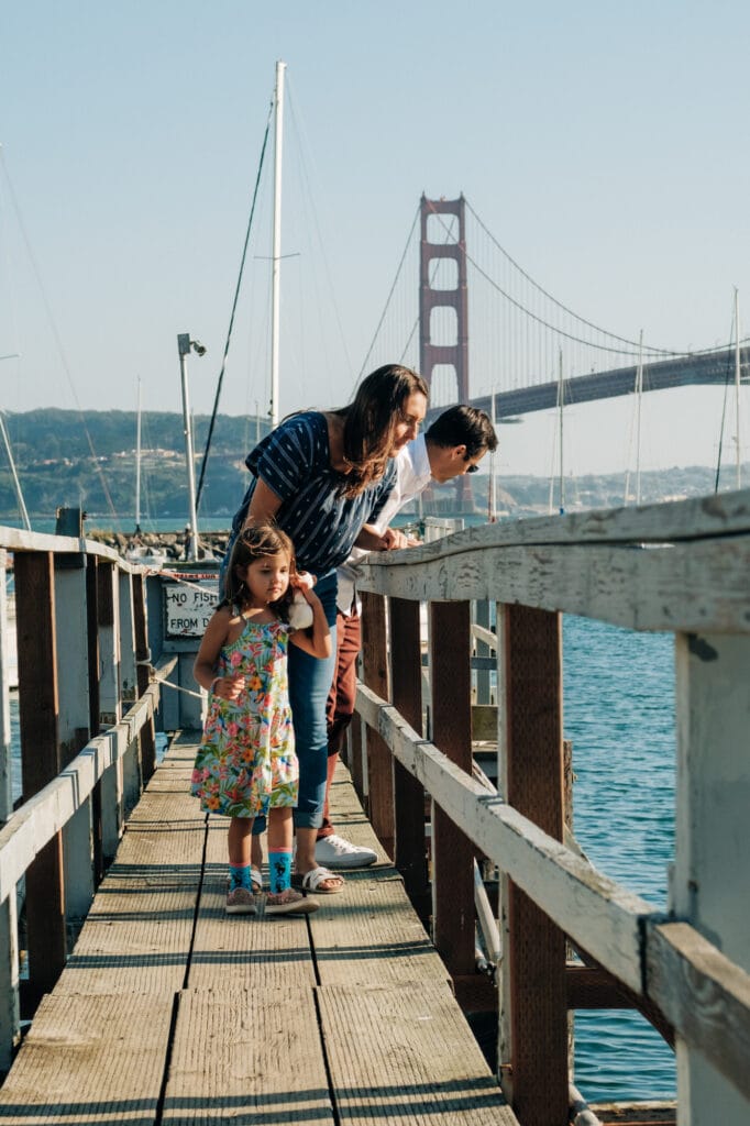 Family of three look into the water off a dock in front of the Golden Gate Bridge.