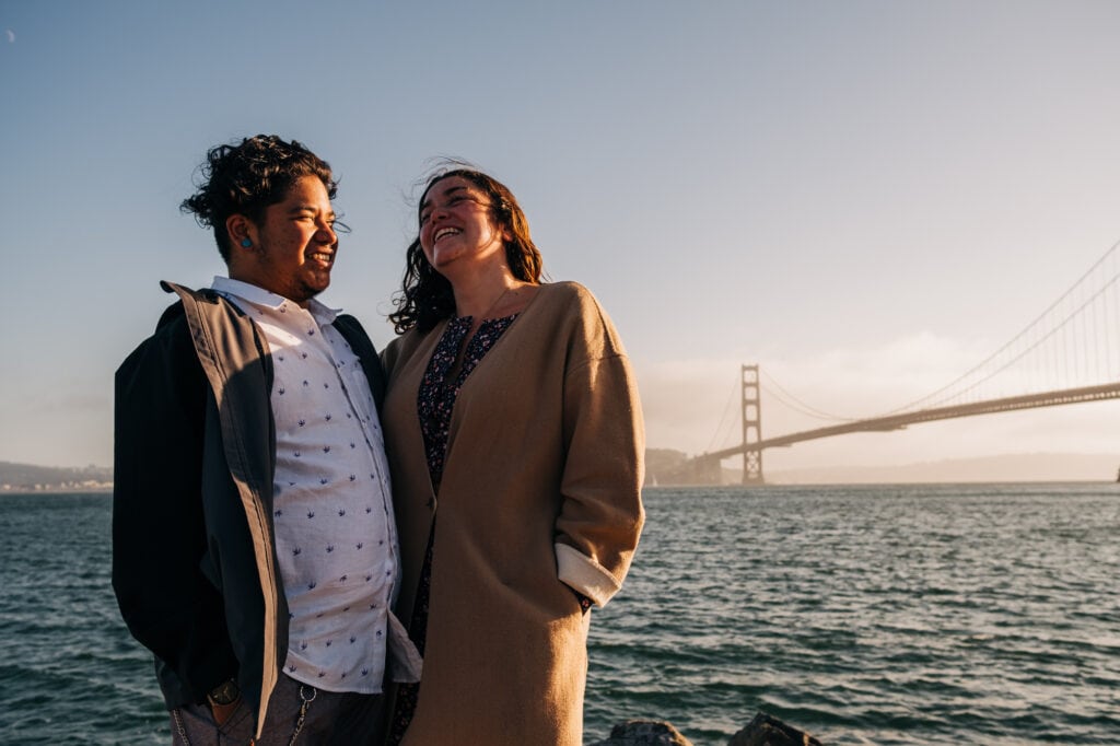 Couple laugh at each other standing on a jetty in front of the Golden Gate Bridge.