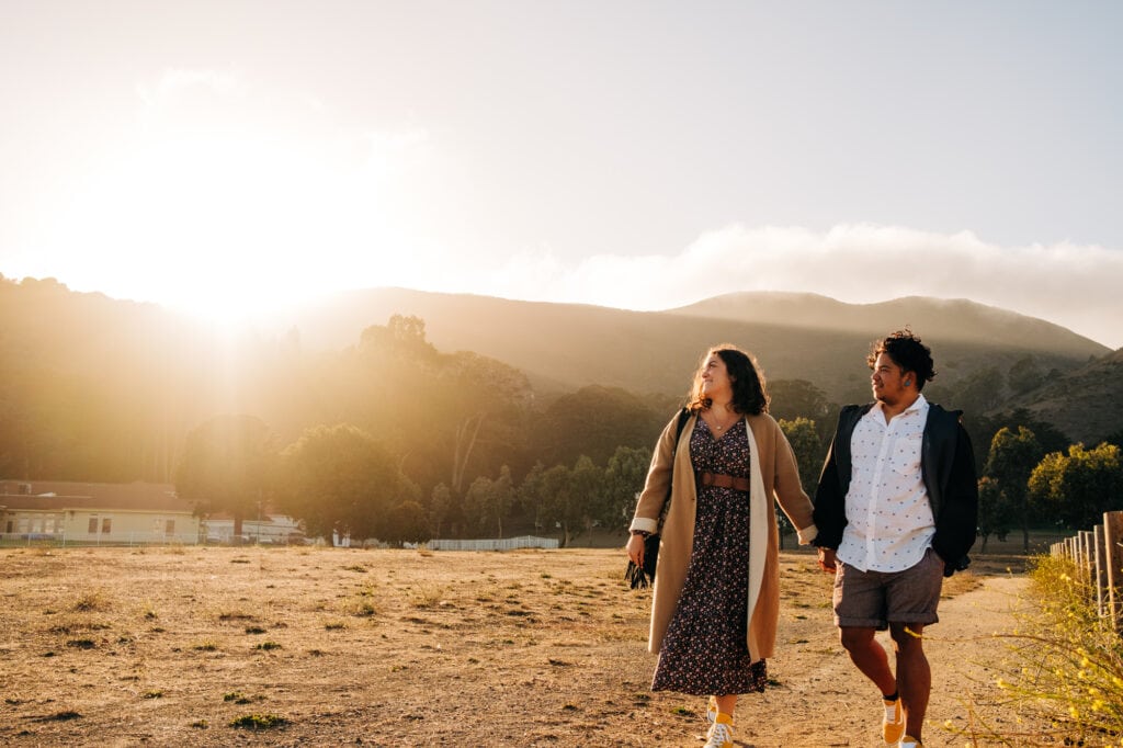 Couple walk hand in hand through a field bathed in California sunset gold light.