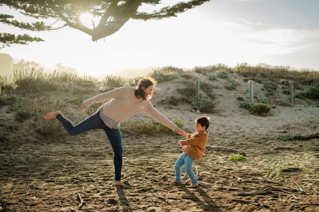 Son pulls mom off balance as they laugh and play under a tree on the beach.