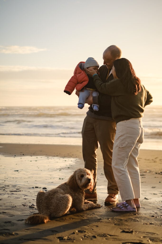 Parents hold infant while dog sits on the sandy beach at their feet.
