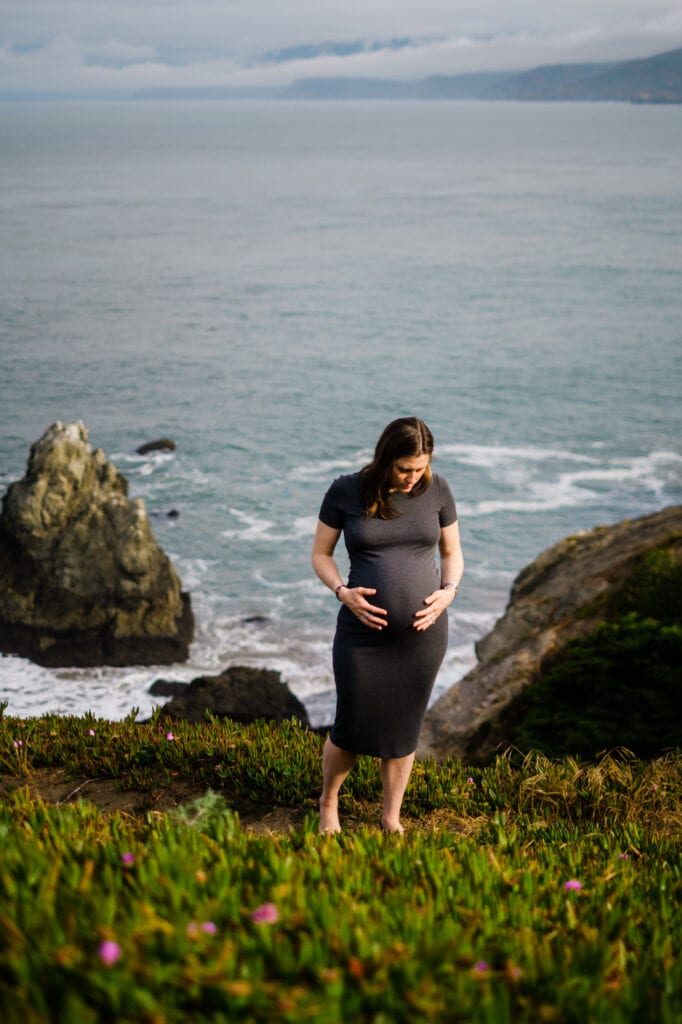 Pregnant woman holding belly while standing above the ocean.