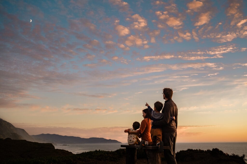 Family of four sits on a bench and looks at the sunset and moonrise.