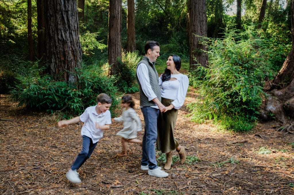 Parents embrace beneath redwoods as kids run circles around them.