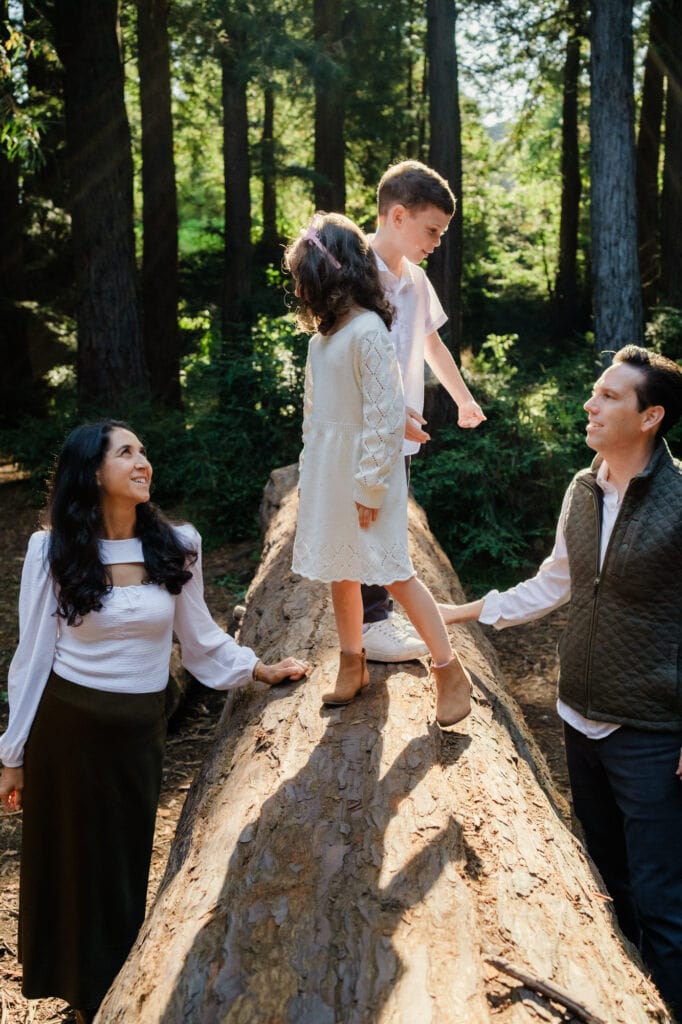 Daughter and son stand on huge fallen log while looking at their parents