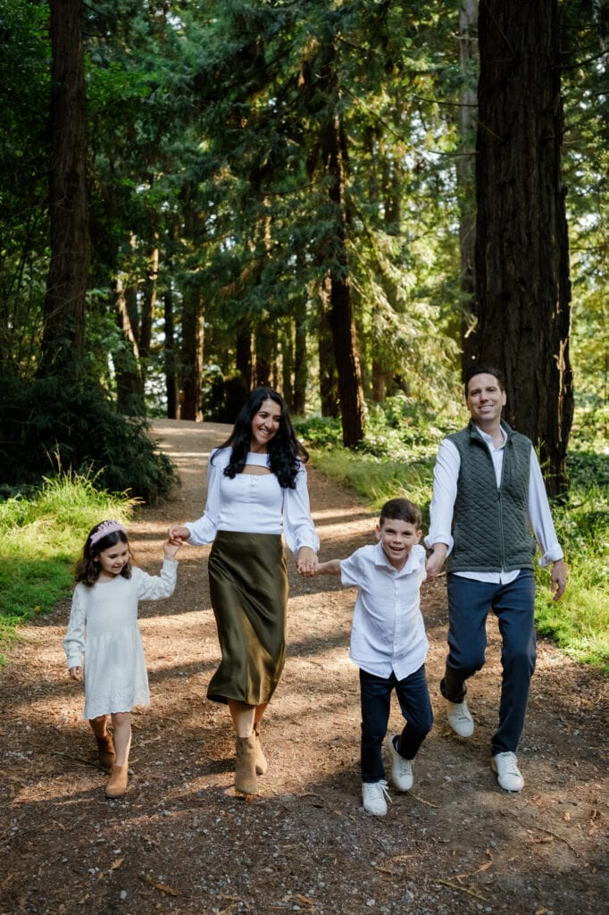 Family holding hands walking down path beneath redwood trees.