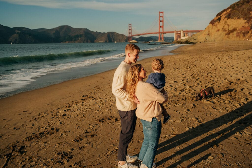 Family of three on the beach look at the Golden Gate Bridge.