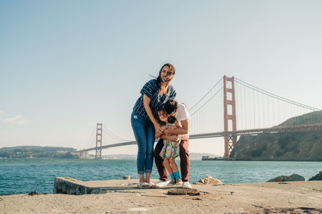 Family of three hug and laugh on a pier in front of the Golden Gate Bridge.