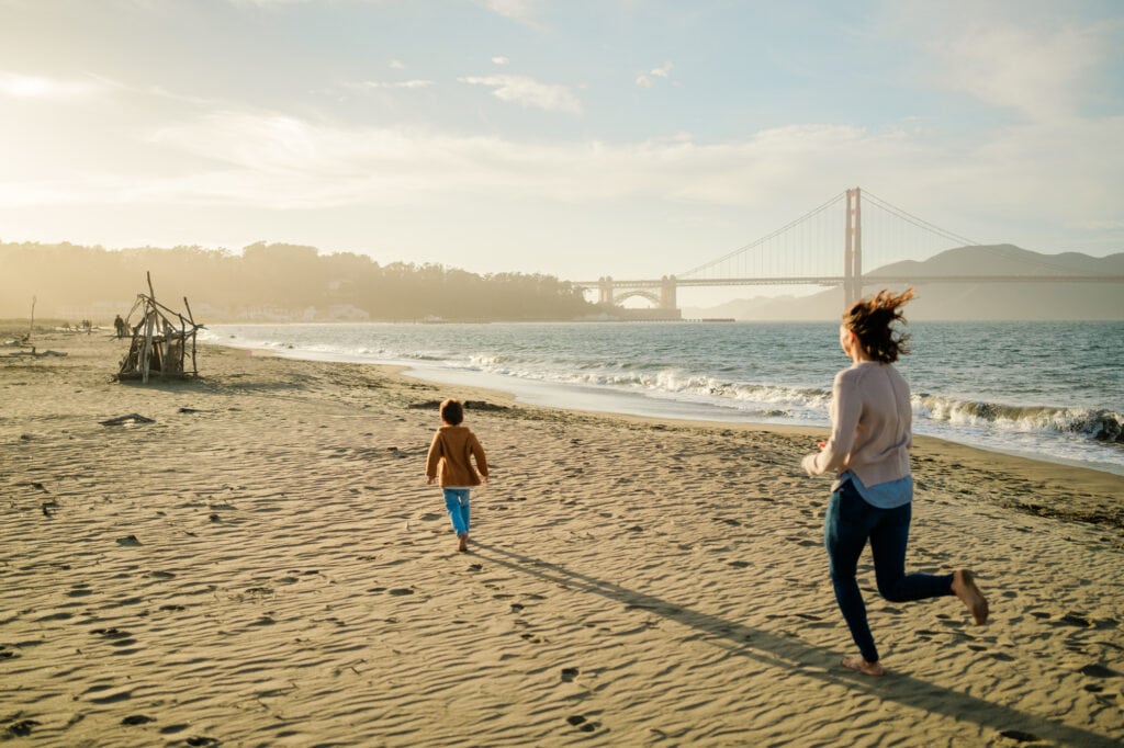 Toddler races down the beach toward the Golden Gate Bridge. Mom sprints to catch up.