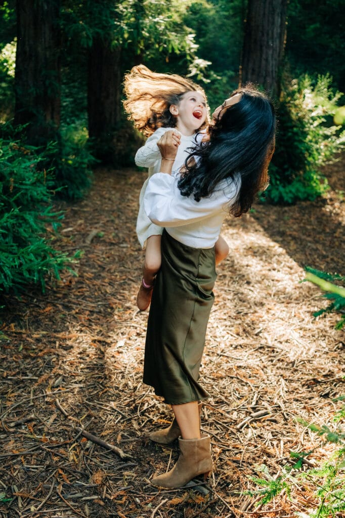 Mom swings daughter while light through the forest illuminates her hair.