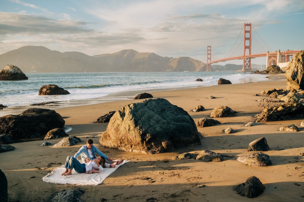 A family sits on a beach blanket with the Golden Gate Bridge in the background.