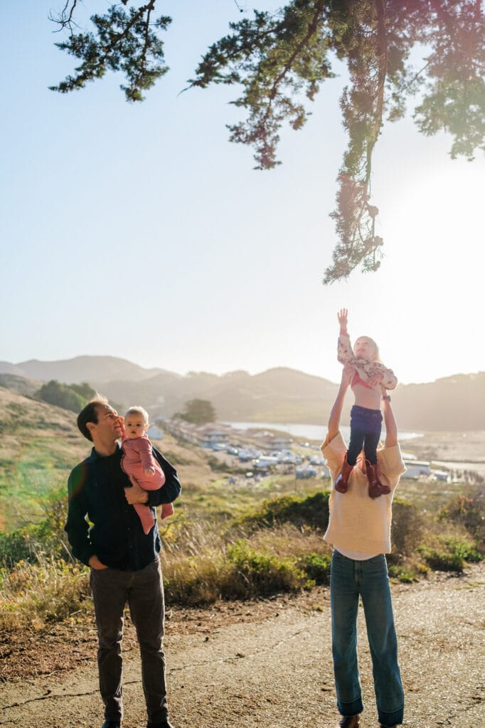 Dad and baby watch as mom lifts daughter to reach for a tree.