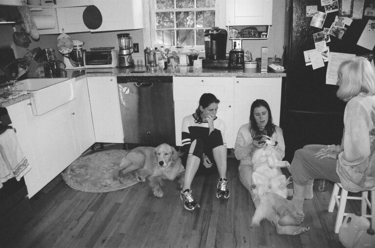 Young woman and her friend sit on the kitchen floor with two dogs while mother looks on.