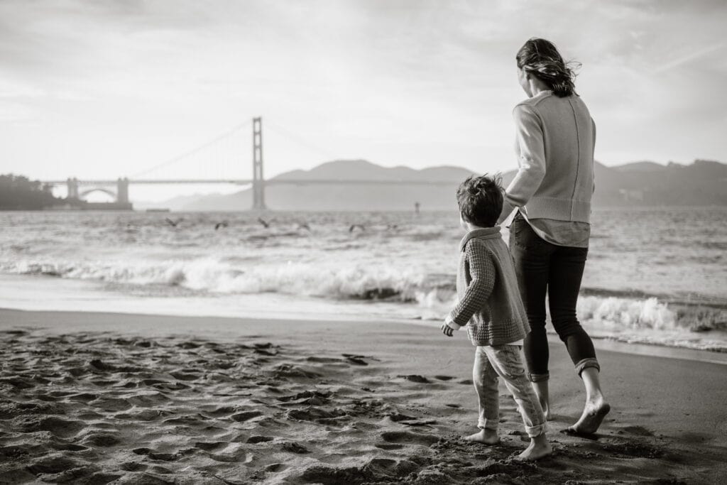 Mom holds young son's hand as they walk up a beach toward the Golden Gate Bridge.