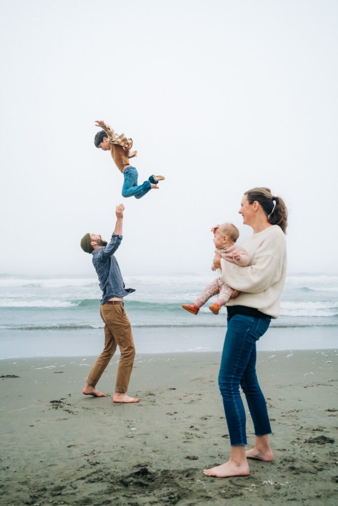 Dad tosses toddler on the beach while mom and baby look on.