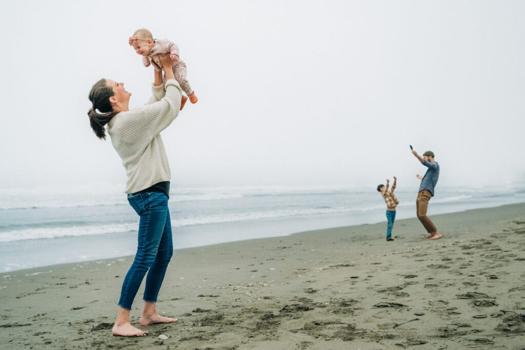 Mom holds baby above head while in the background dad dances with toddler on the beach in the fog.