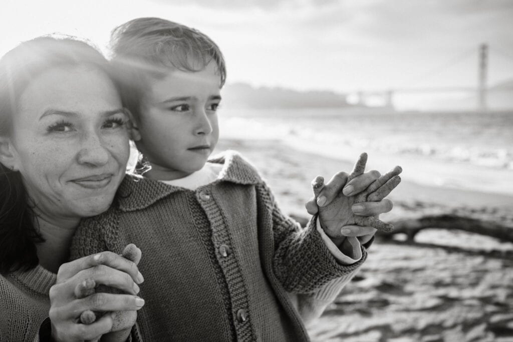 Black and white of mom clasping son's hands as sunset light streams over her shoulder.