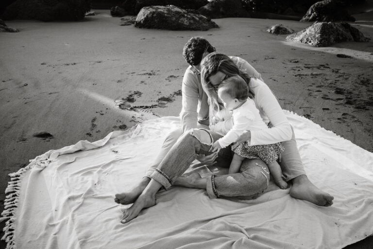 Black and white with family of three embracing tightly while sitting on a beach blanket in a sliver of sunset light.