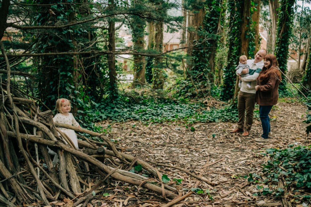 Mom, dad, and baby look on as toddler daughter explores little house made of sticks.