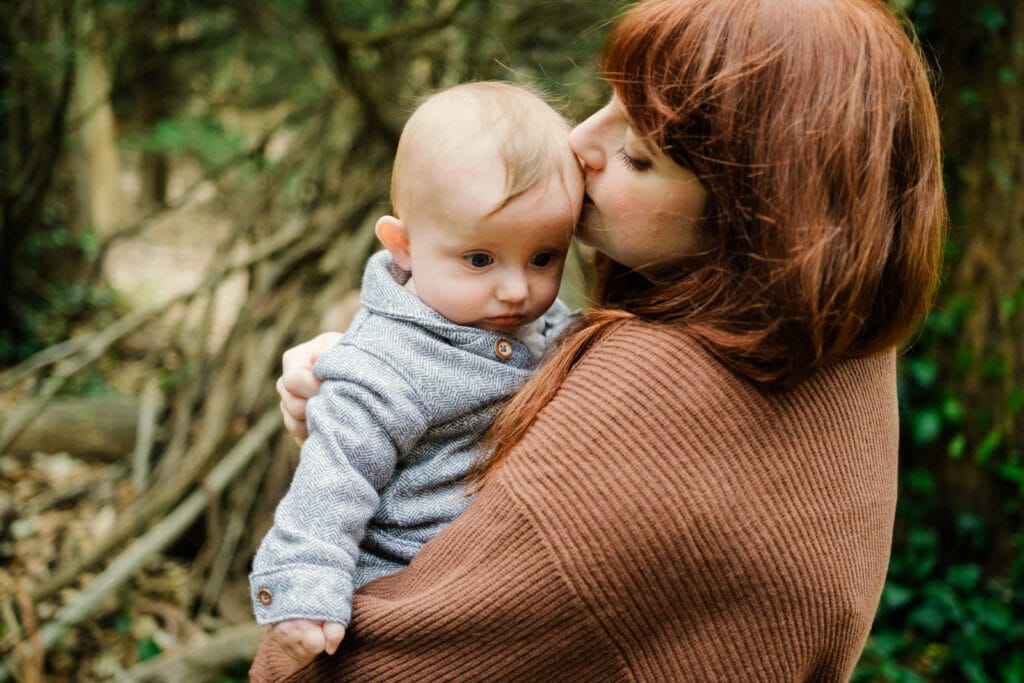 Mom tenderly kisses newborn's head while standing in a foggy forest.