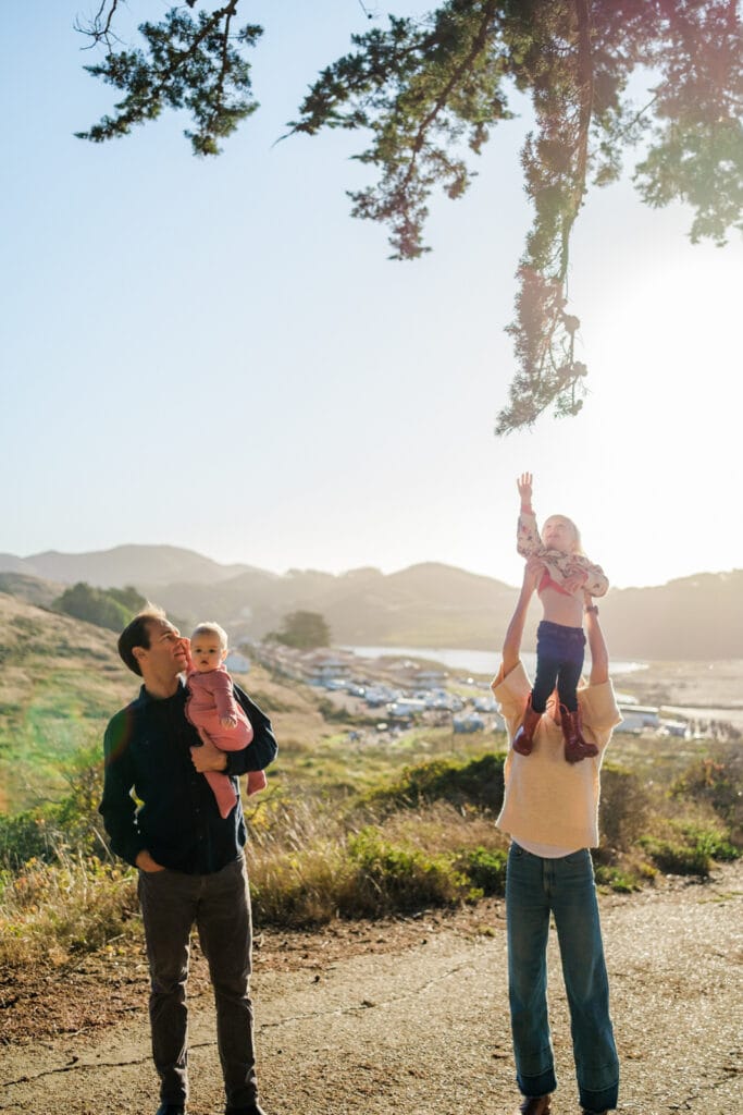 Mom holds daughter up to reach tree with iconic Fort Cronkite buildings in the background.