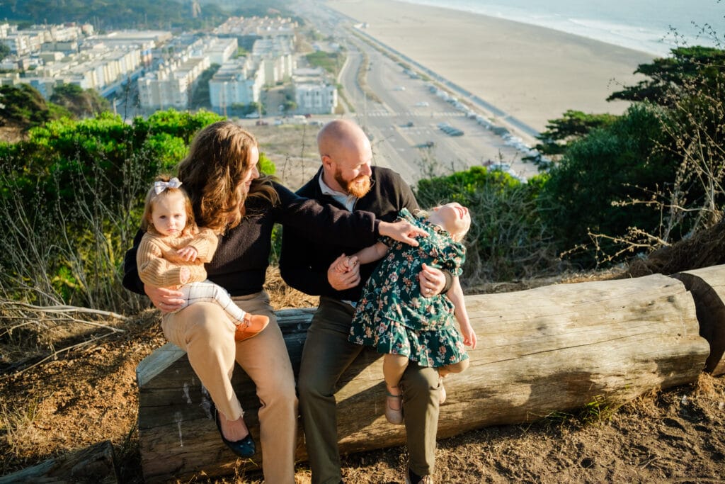 Family with baby and preschooler sit on a log overlooking the beach and the ocean.