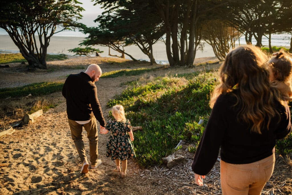 Dad holds daughter's hand as they walk down a sandy path to a level spot under coastal trees.