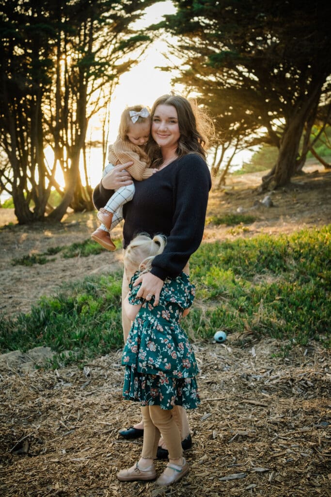 Mom holds baby daughter on hip while holding her preschooler close with the other hand. Sunset light through the trees.