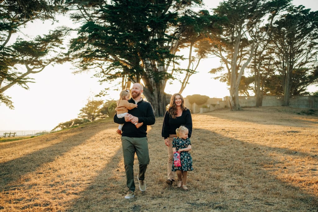Family of four walk together with the sun just blocked behind them by a towering Monterey Pine.