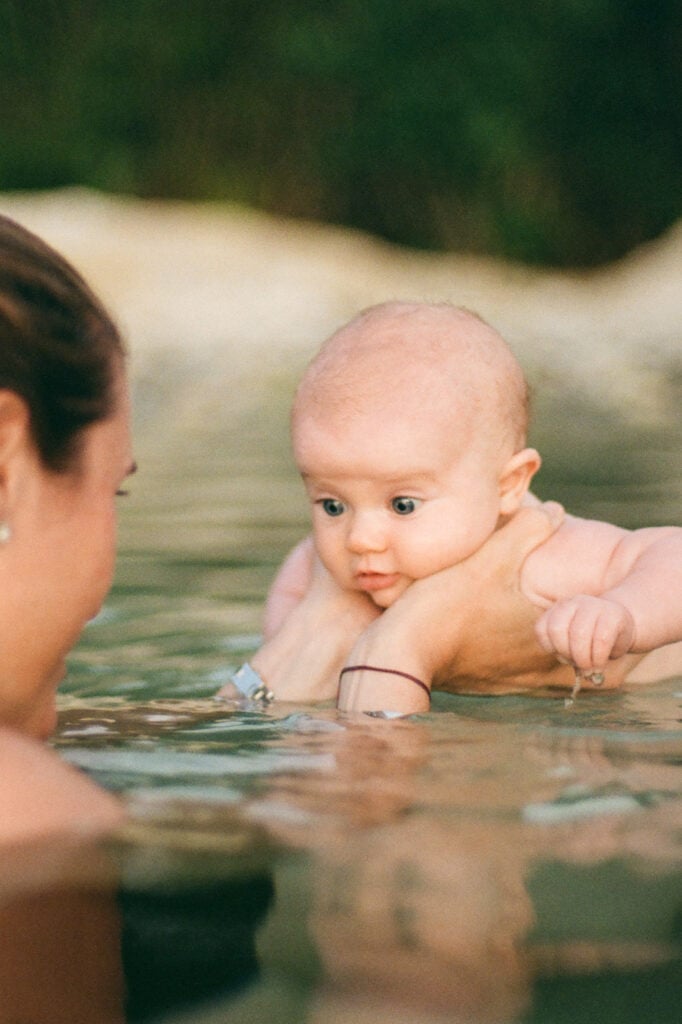 Mom holds baby daughter in tropical ocean as she learns to swim.