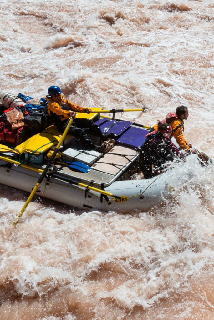 Man rows raft through big whitewater with two passengers on the bow.