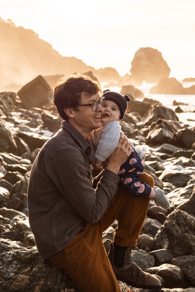Man squeezes baby to cheek in front of sunset and rocky beach.