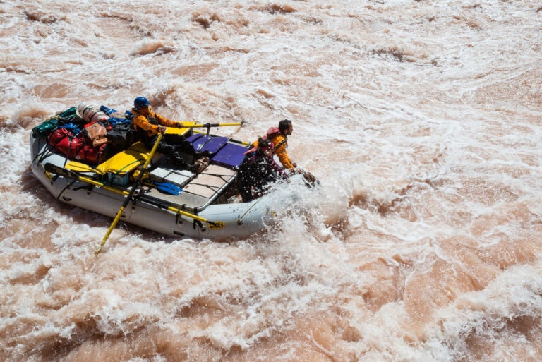 Man rows whitewater raft through huge rapid while crew sits on the bow.